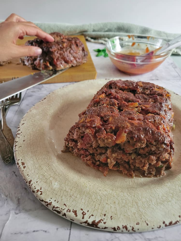 meatloaf on a plate, more meatloaf in the background on a cutting board with a hand and a knife, next to a bowl of bbq sauce