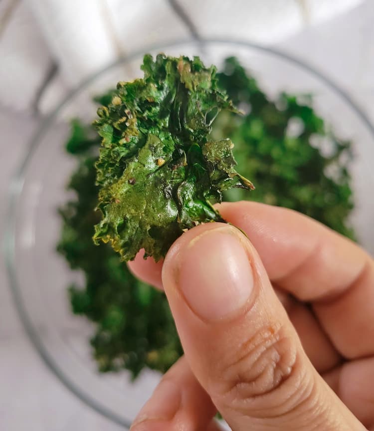 hand holding a kale chip over a bowl of the rest