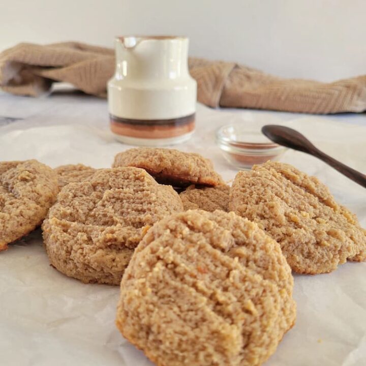 pile of almond flour cookies on a piece of parchment paper, small ramekin of cinnamon and a spoon in the background next to a milk pitcher