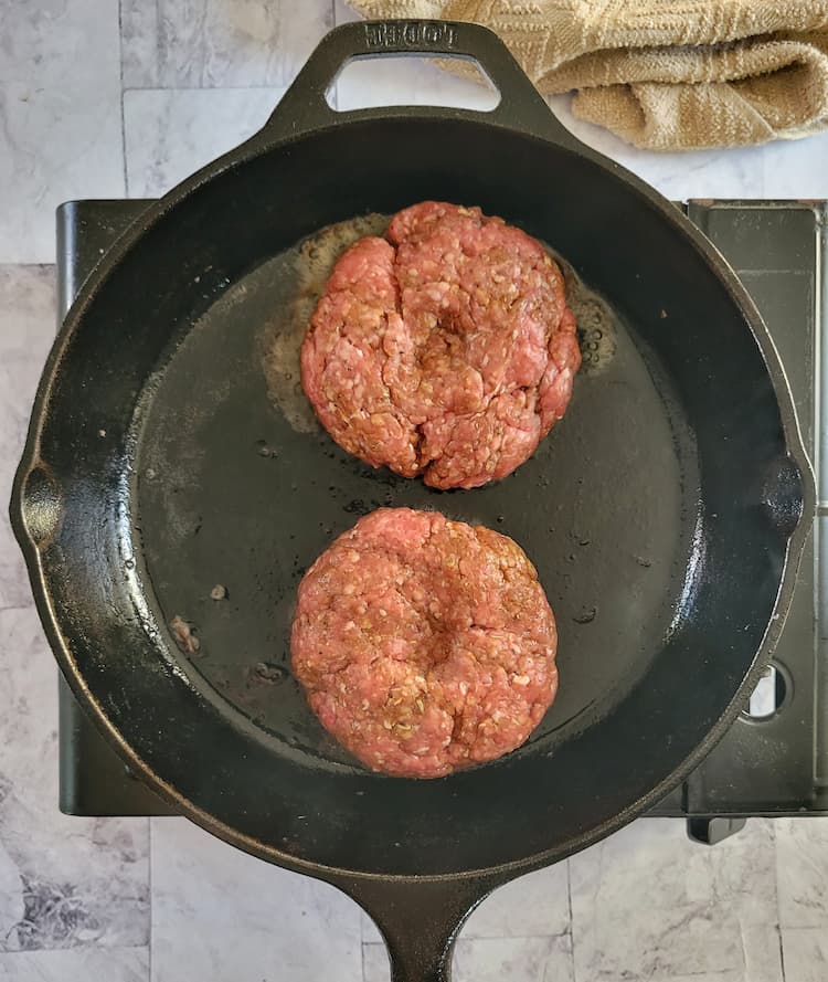 black cast iron skillet with two round beef patties with thumbprints in the centers