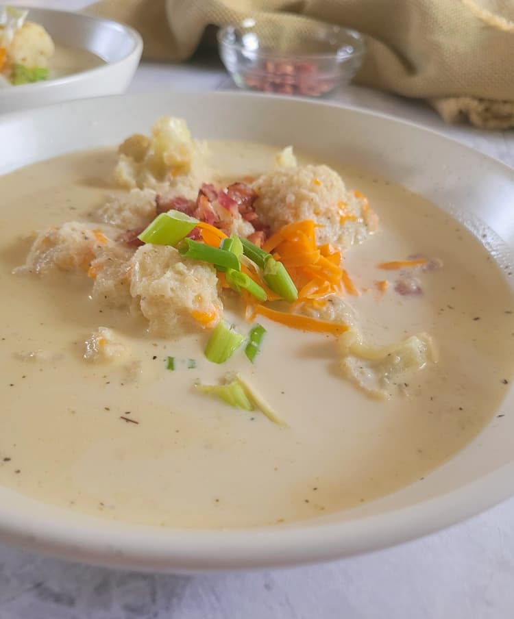 side view of of a bowl of keto cauliflower soup topped with bacon, green onions and cheddar cheese, another bowl of soup in the background next to a bowl of bacon