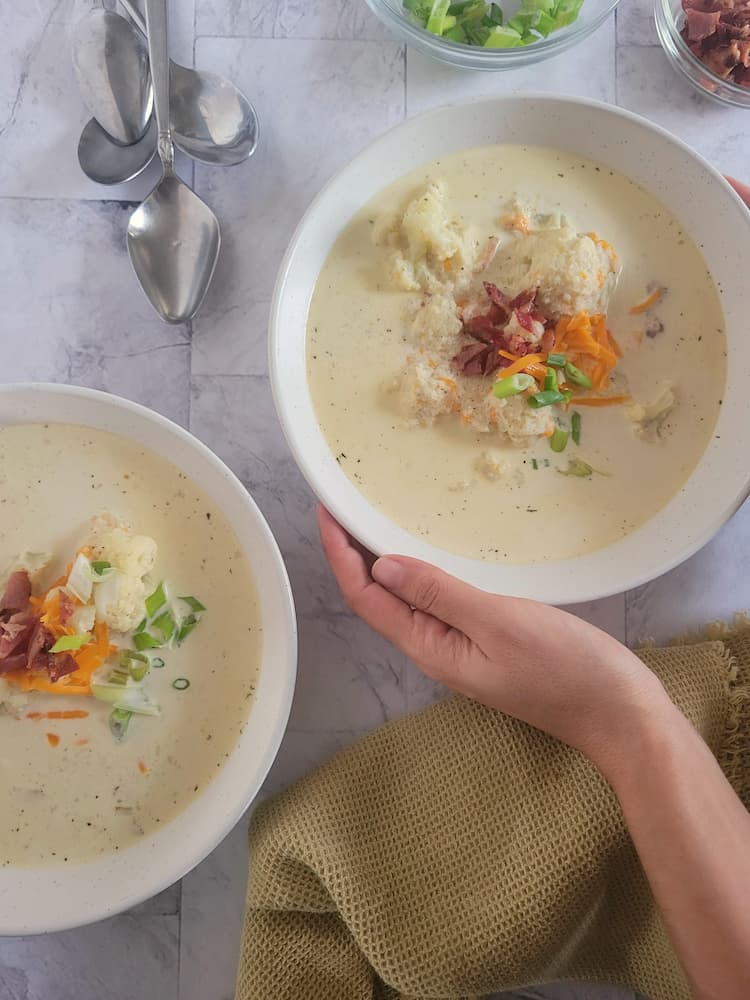 two bowls of keto cauliflower soup topped with bacon, grated cheddar cheese and green onions, hands holding one bowl, 4 spoons in the background next to bowls of green onions and bacon