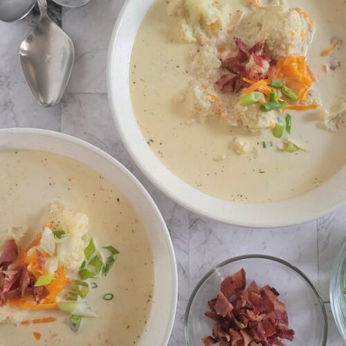 two bowls of keto cauliflower soup topped with bacon, grated cheddar cheese and green onions next to a bowl of bacon and 4 spoons