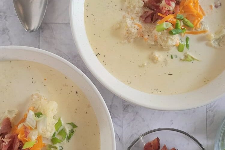 two bowls of keto cauliflower soup topped with bacon, grated cheddar cheese and green onions next to a bowl of bacon and 4 spoons