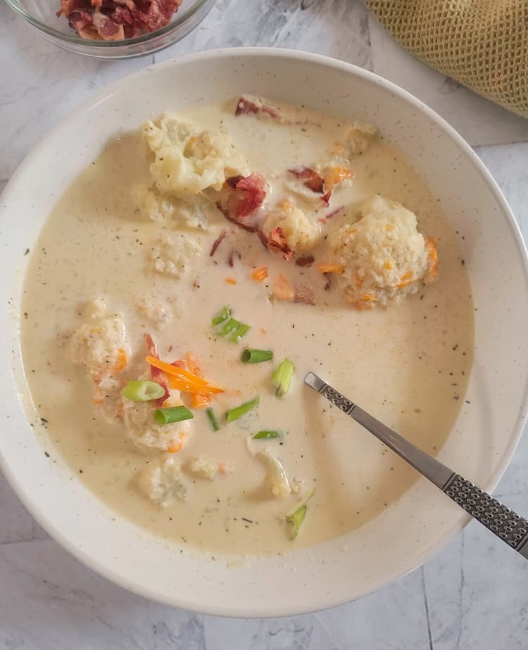 big bowl of keto cauliflower soup with florets, green onion, bacon and cheddar cheese, bowl of bacon in the background, spoon in the bowl