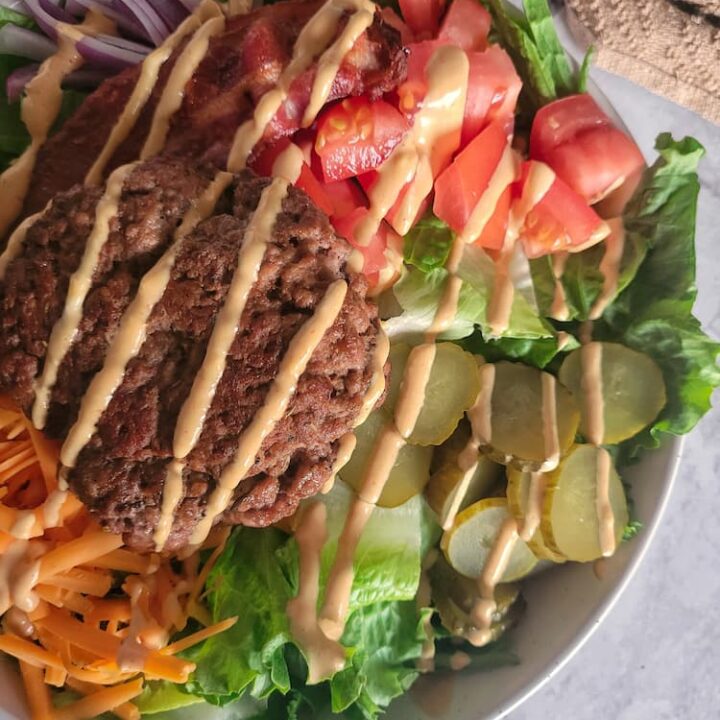 bowl of lettuce, chopped pickles, diced tomatoes, grated cheddar cheese, bacon, sliced red onion, and a burger patty and a creamy orange sauce drizzled on top