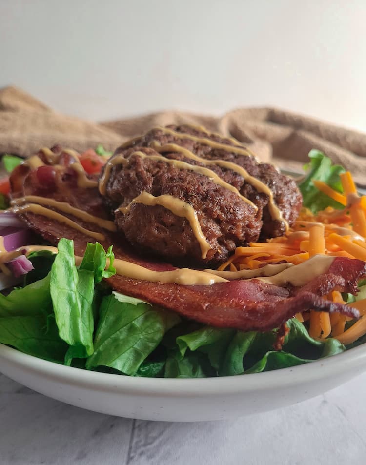 side view of a burger bowl drizzle with creamy orange sauce, lettuce, bacon, cheddar cheese and a beef burger patty visible