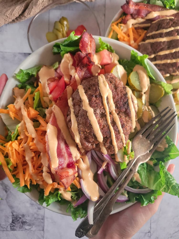 bowl of lettuce, chopped pickles, diced tomatoes, grated cheddar cheese, bacon, sliced red onion, and a burger patty and a creamy orange sauce drizzled on top. Another bowl of the same thing in the background next to a bowl of pickles and tomatoes