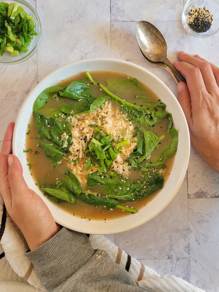 hand on a bowl of egg drop soup with egg, spinach and sesame seeds, other hand on spoons, green onions in the background with sesame seeds