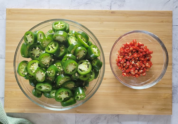 a bowl of sliced jalapeños next to a bowl of thai red chilies on a cutting board