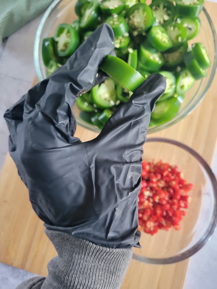 hand with a black glove holding a jalapeño slice over a bowl with the rest, another bowl of sliced thai red chilies beside it