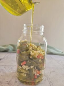 olive oil being poured into a mason jar with sliced jalapeños, thai red chilies and minced garlic