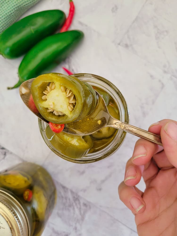 hand holding a spoon with some jalapeños and thai chilies on it over a jar with the rest, more peppers in the background