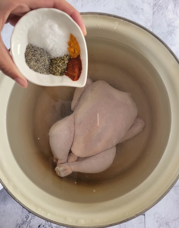 hand holding a ramekin of different spices about to be poured into a pot of water with a whole chicken