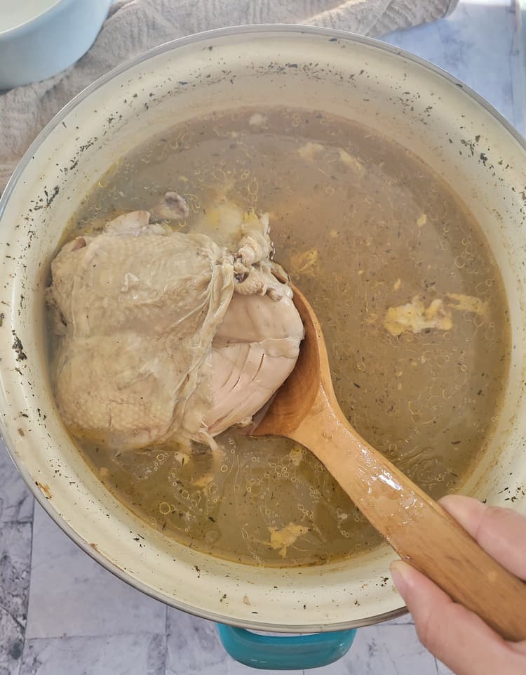 hand with a wooden spoon lifting some cooked chicken out of a pot of broth