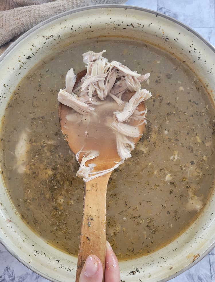 hand holding a wooden spoon with shredded chicken over a pot of brothy soup