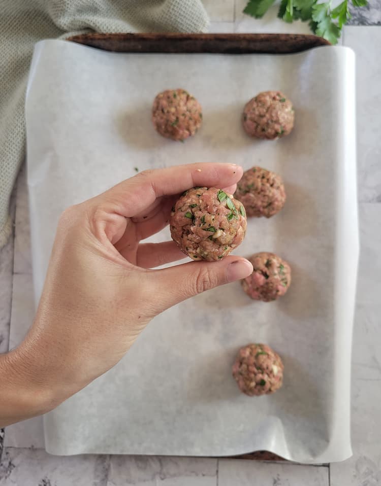 hand holding a raw meatball over a parchment lined baking sheet with the rest