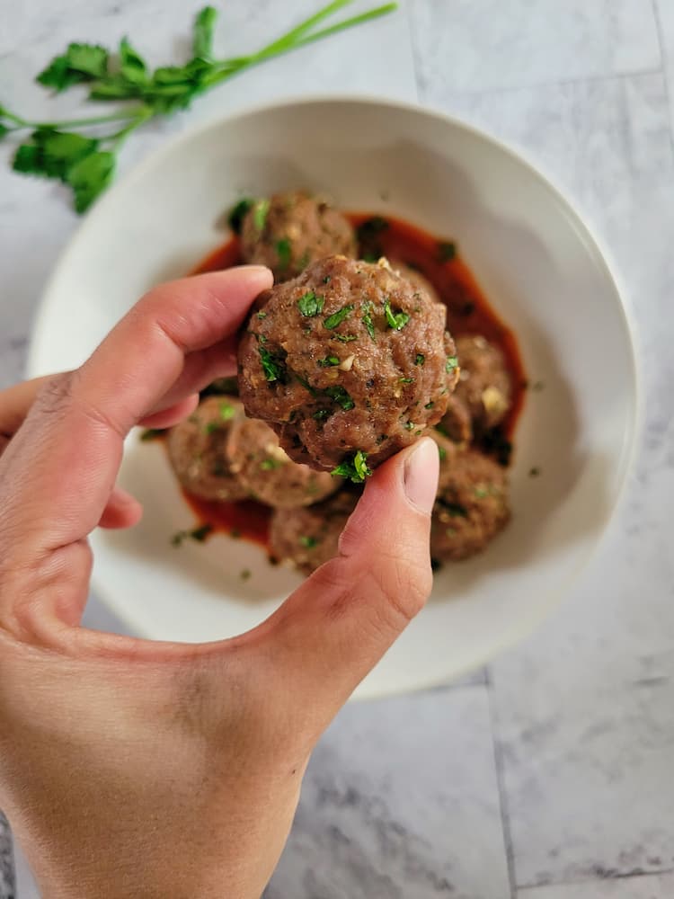 hand holding a cooked meatball over a bowl with the rest