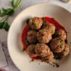 a bowl of gluten free meatballs garnished with parsley, bowl of sauce and fresh herbs in the background