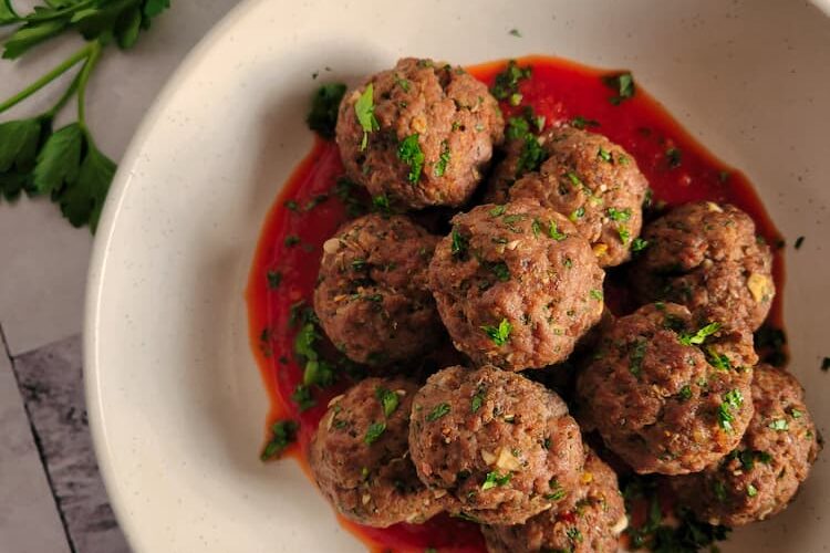 a bowl of gluten free meatballs garnished with parsley, bowl of sauce and fresh herbs in the background