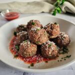 bowl of gluten free meatballs sitting in tomato sauce, garnished with fresh chopped parsley and shaved parmesan cheese, ramekin of tomato sauce in the background