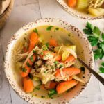 bowl of no noodle chicken soup with chopped parsley, baby carrots, celery and shredded chicken, spoon in the bowl, another bowl in the background