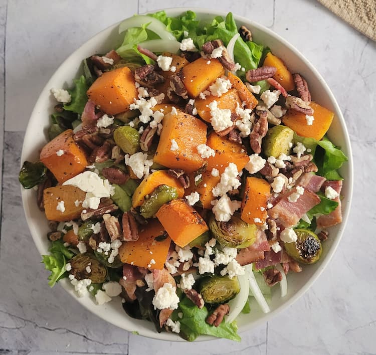 bowl of greens with cubed butternut squash, halved brussels sprouts, pecans, feta and onions