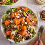 bowl of greens with cubed butternut squash, halved brussels sprouts, pecans, feta and onions, two forks next to the bowl, hand on the forks, another salad in the background next to a bowl of creamy dressing and ramekin of pecans