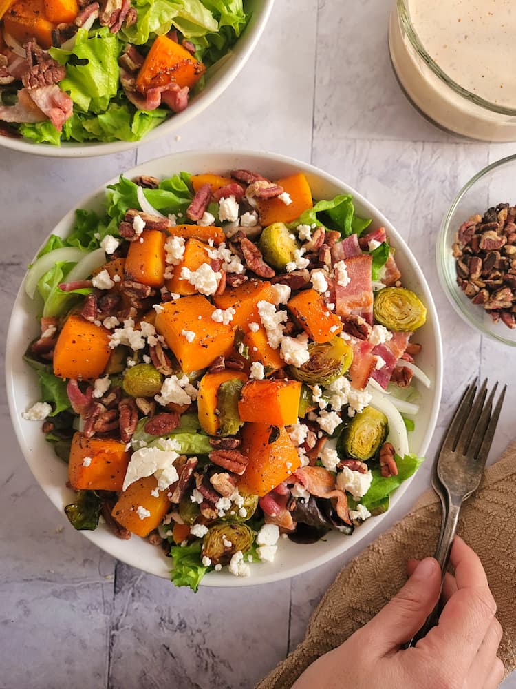 bowl of greens with cubed butternut squash, halved brussels sprouts, pecans, feta and onions, two forks next to the bowl, hand on the forks, another salad in the background next to a bowl of creamy dressing and ramekin of pecans