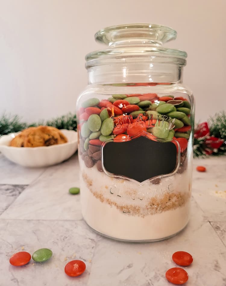jar of christmas smarties, chocolate chips, flour and sugars, christmas garland with red bows in the background with a bowl of cookies and scattered smarties