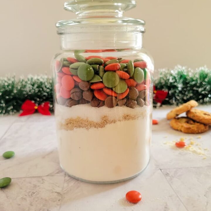 jar of christmas smarties, chocolate chips, flour and sugars, christmas garland with red bows in the background with 3 cookies and scattered smarties