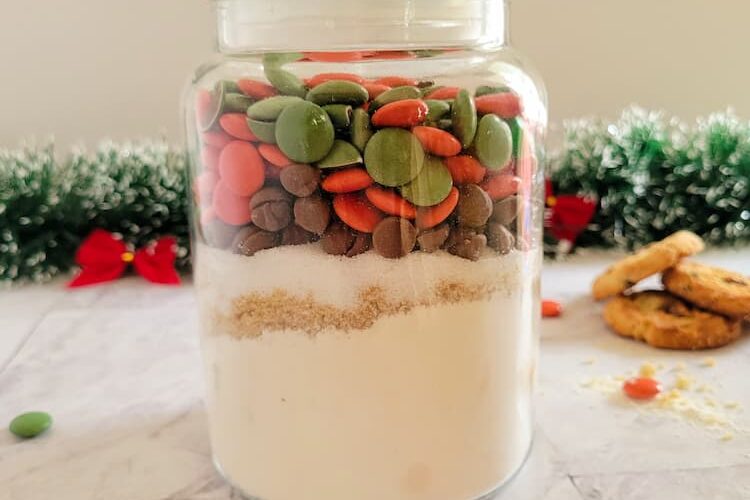 jar of christmas smarties, chocolate chips, flour and sugars, christmas garland with red bows in the background with 3 cookies and scattered smarties