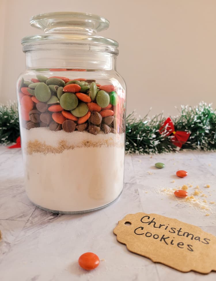 jar of christmas smarties, chocolate chips, flour and sugars, christmas garland with red bows in the background with scattered smarties and a cardboard label that says christmas cookies