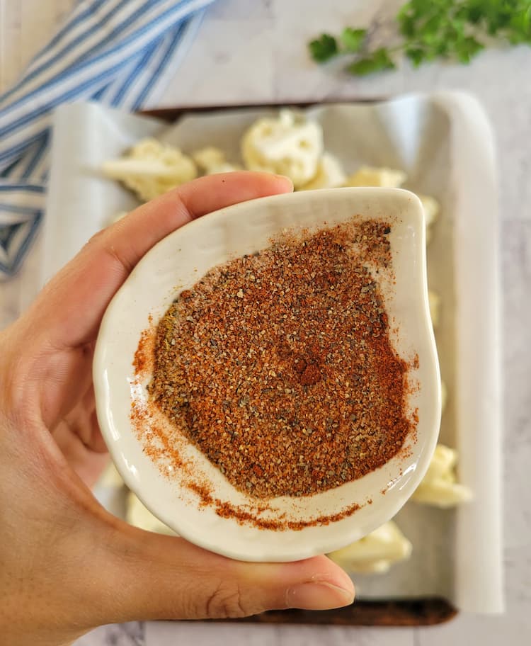 hand holding a small ramekin of mixed spices over a sheet pan of cauliflower florets