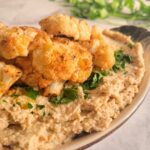 side view of a dish with cauliflower hummus topped with chopped parsley and roasted and seasoned florets, more parsley in the background