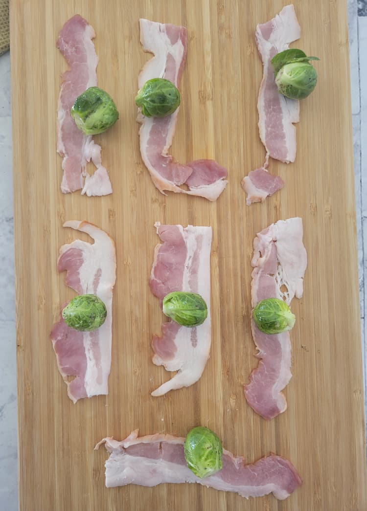 7 half strips of raw bacon on a cutting board with a whole brussels sprout in the middle of each