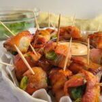 tray of bacon wrapped brussels sprouts with toothpicks, orange dipping sauce in the middle, bowl of brussels sprouts in the background