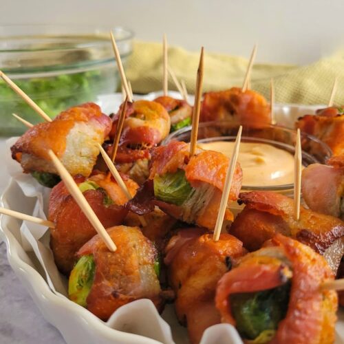tray of bacon wrapped brussels sprouts with toothpicks, orange dipping sauce in the middle, bowl of brussels sprouts in the background