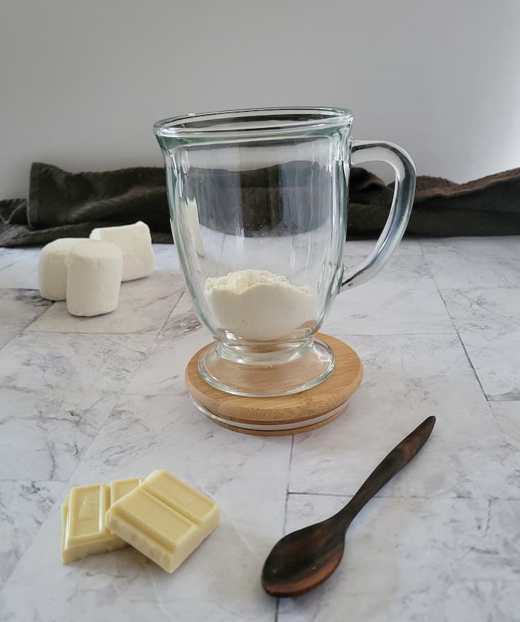 glass mug on a brown coaster with white powder inside, 3 large marshmallows, two pieces of white chocolate and a wooden spoon in the background
