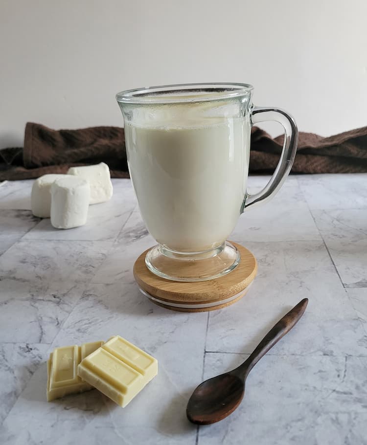 glass mug with white hot chocolate on a brown coaster, marshmallows in the background, spoon and 2 pieces of white chocolate in the front
