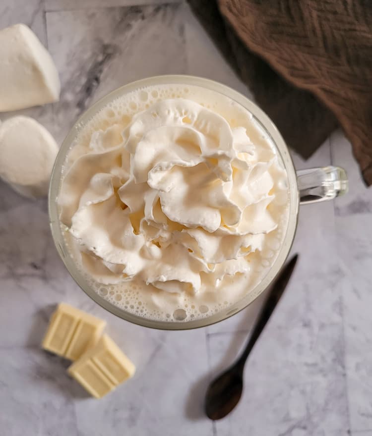 whipped cream topped glass mug, marshmallows, spoon and pieces of white chocolate in the background