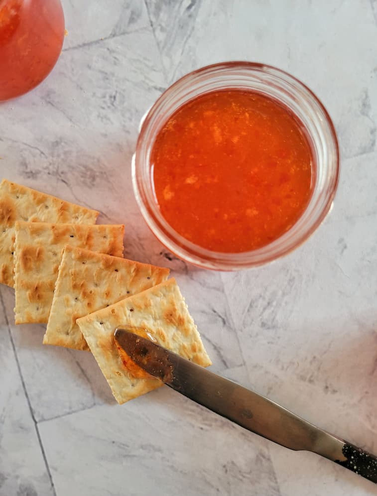 4 saltine crackers and a knife with some red pepper jelly in a jar and on one of the crackers