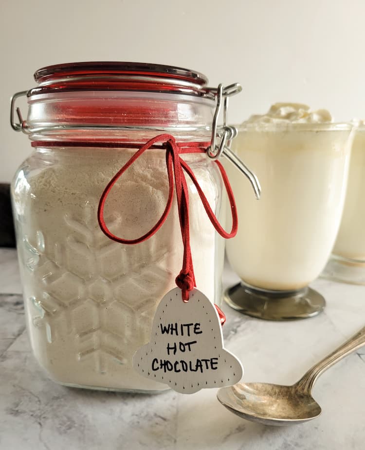 jar of homemade white hot chocolate mix with a label, mugs of white hot chocolate in the background with a spoon