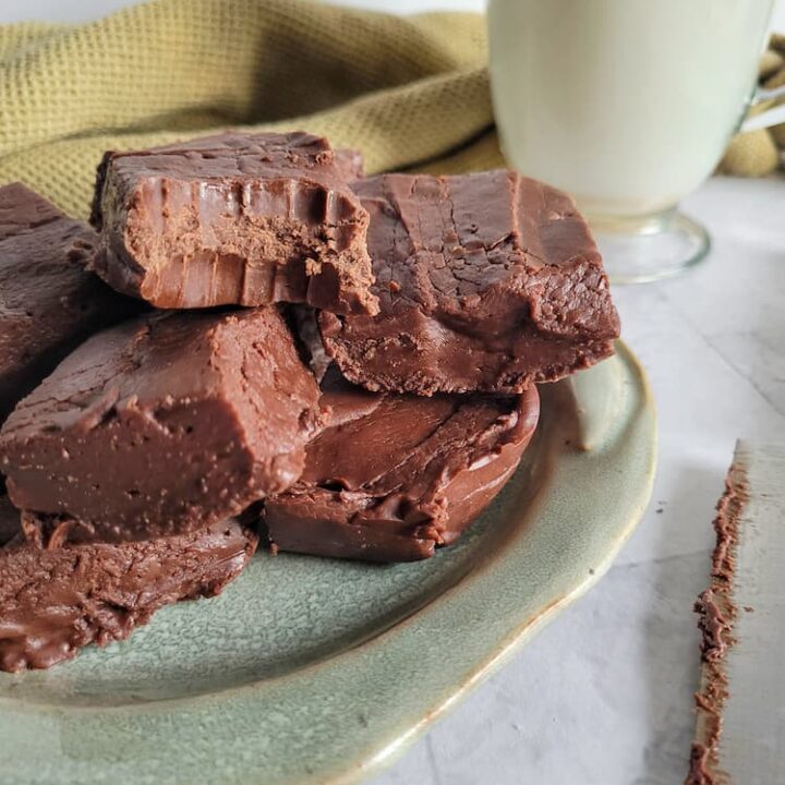 pieces of chocolate fudge piled on a plate, glass of milk in the background with a knife, bite marks in one of the pieces of fudge