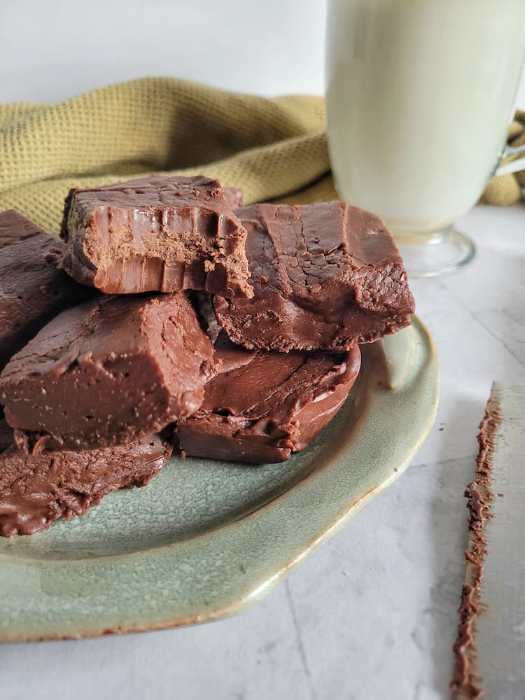 pieces of chocolate fudge piled on a plate, glass of milk in the background with a knife, bite marks in one of the pieces of fudge