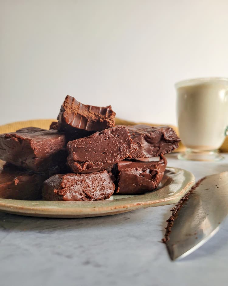 pieces of chocolate fudge piled on a plate, glass of milk in the background with a knife, bite marks in one of the pieces of fudge