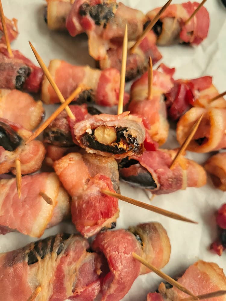 pile of bacon wrapped dates with toothpicks, one of the dates is bitten showcasing the almond in the middle
