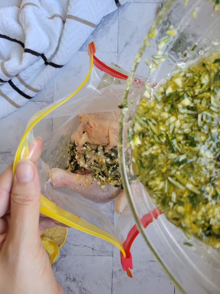 hand holding a ziploc bag open with chicken pieces, bowl of oil, herbs, garlic and spices being poured on top