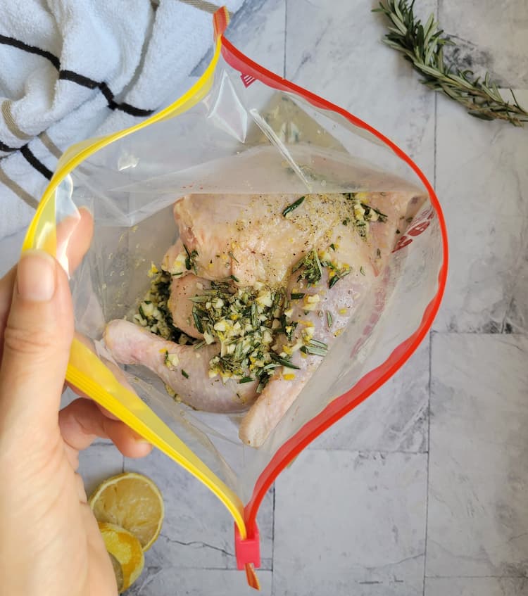 hand holding a ziploc bag open with chicken leg quarters and herbs, garlic and seasonings on top