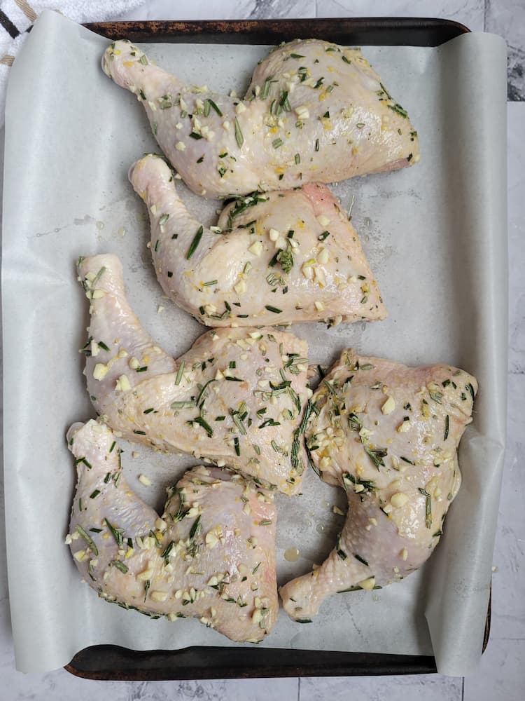 parchment lined baking sheet with raw chicken leg quarters seasoned with fresh rosemary, minced garlic and spices